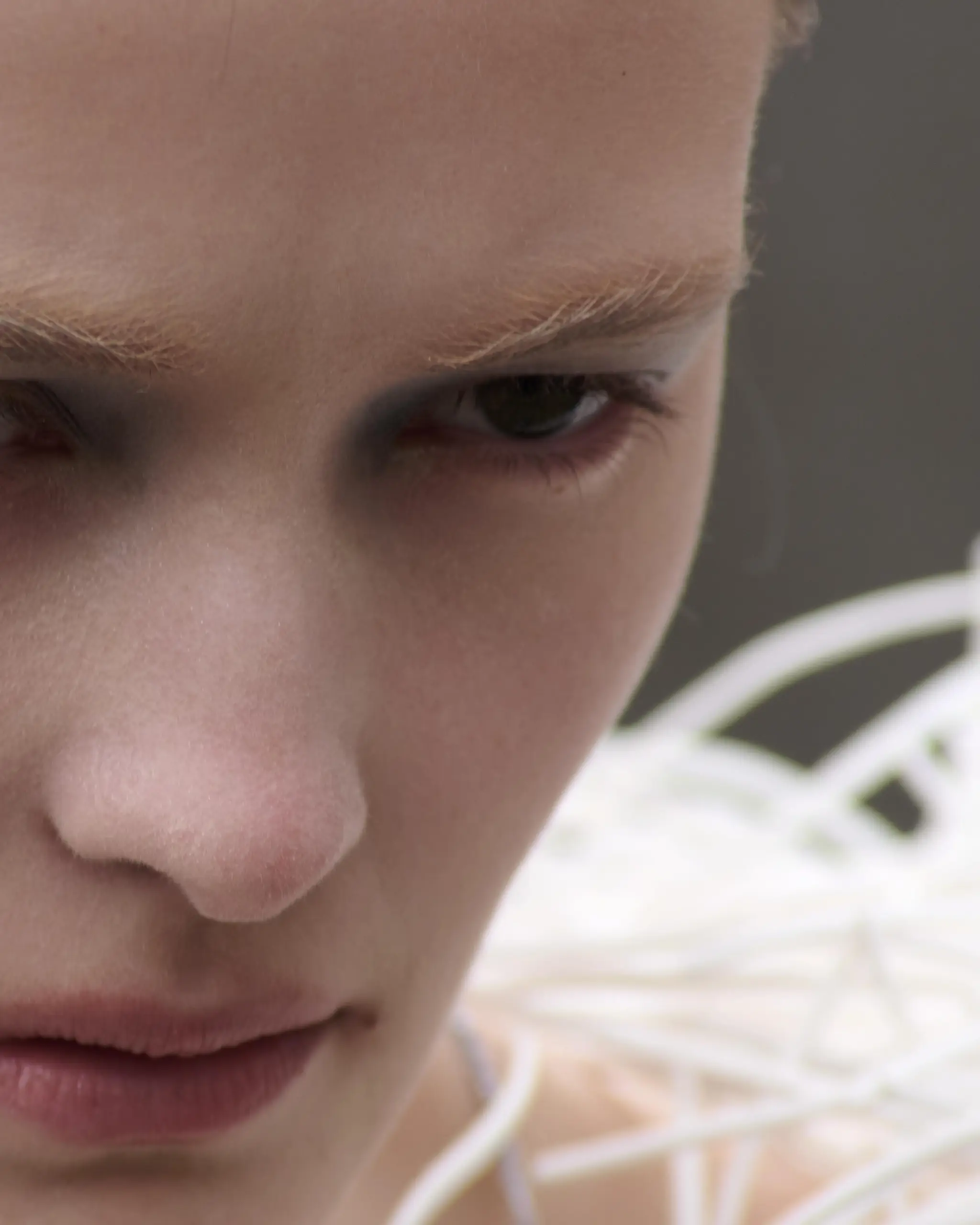 Extreme close-up of a face with pale brows and a downward gaze, tangled white cord resting against the cheek.