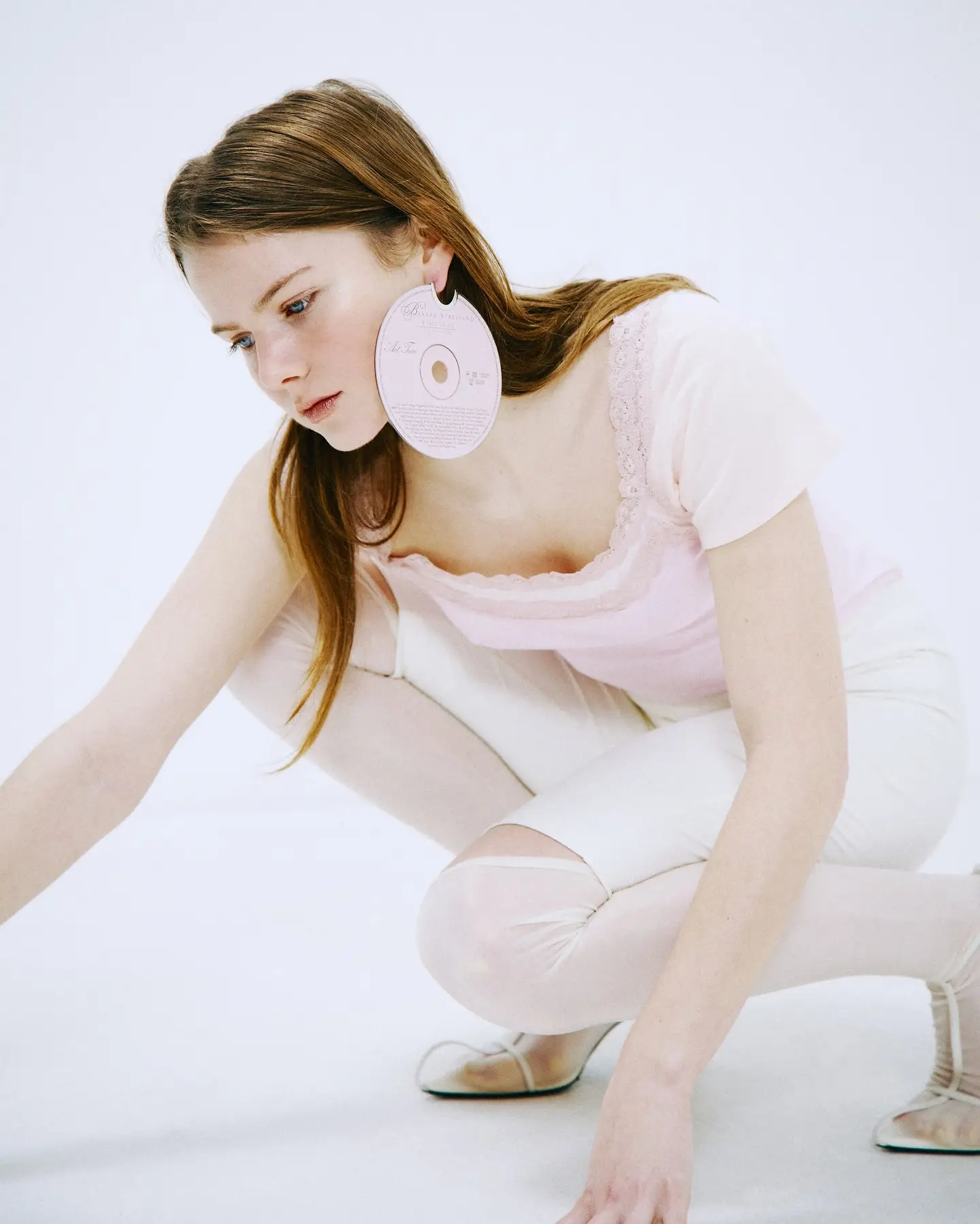 A model crouching on a pale cyclorama in layered cream and pink knitwear, a compact disc worn as an earring.